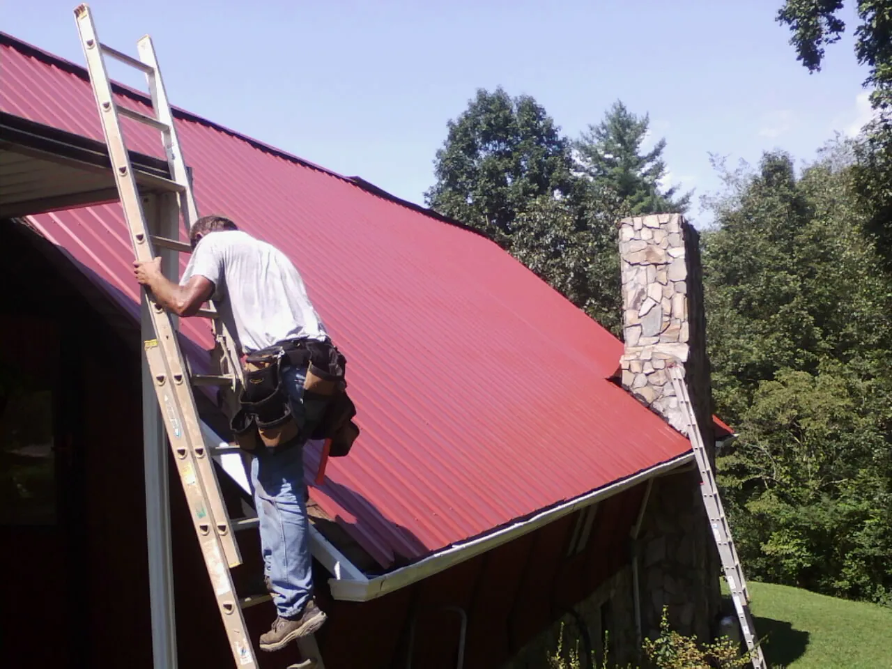 Roofer on a ladder beside a red metal roof and stone chimney — Soffit & Fascia Repair by Roofing Brothers in Richland Center